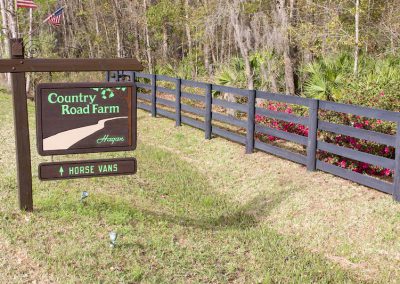 Decretive wooden sign with Country Road Farm name on it hanging from a wood post outside next to ranch style fence and trees in the background.